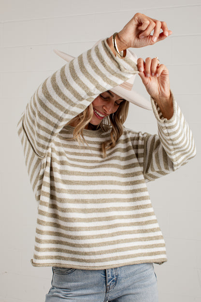 A woman wearing a beige and cream horizontally striped knit sweater, light blue jeans, and a wide-brimmed straw hat, posing with her hands near her head against a white paneled wall.