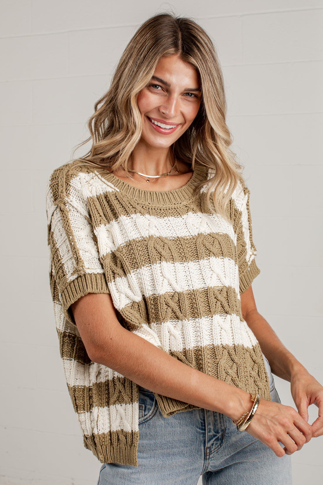 A woman wearing a short-sleeve knit sweater with horizontal stripes in tan and off-white, paired with light blue jeans, against a light gray paneled wall.