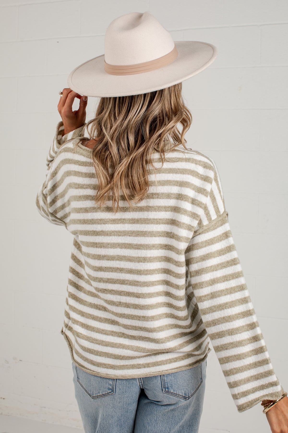A woman shown from the back wearing a cream and tan striped knit sweater, a light beige wide-brim hat, and light blue jeans against a white paneled wall.