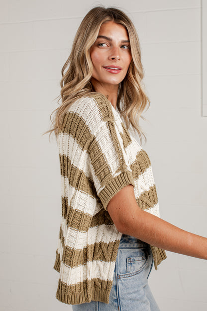 A woman wearing a chunky knit short-sleeve sweater with horizontal stripes in beige and off-white, paired with light-wash blue jeans, standing against a white paneled wall.