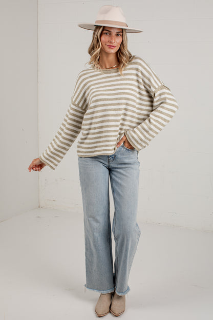 A woman modeling a beige and white striped knit sweater, paired with light-wash wide-leg jeans, beige ankle boots, and a wide-brimmed beige hat, standing against a white studio wall.