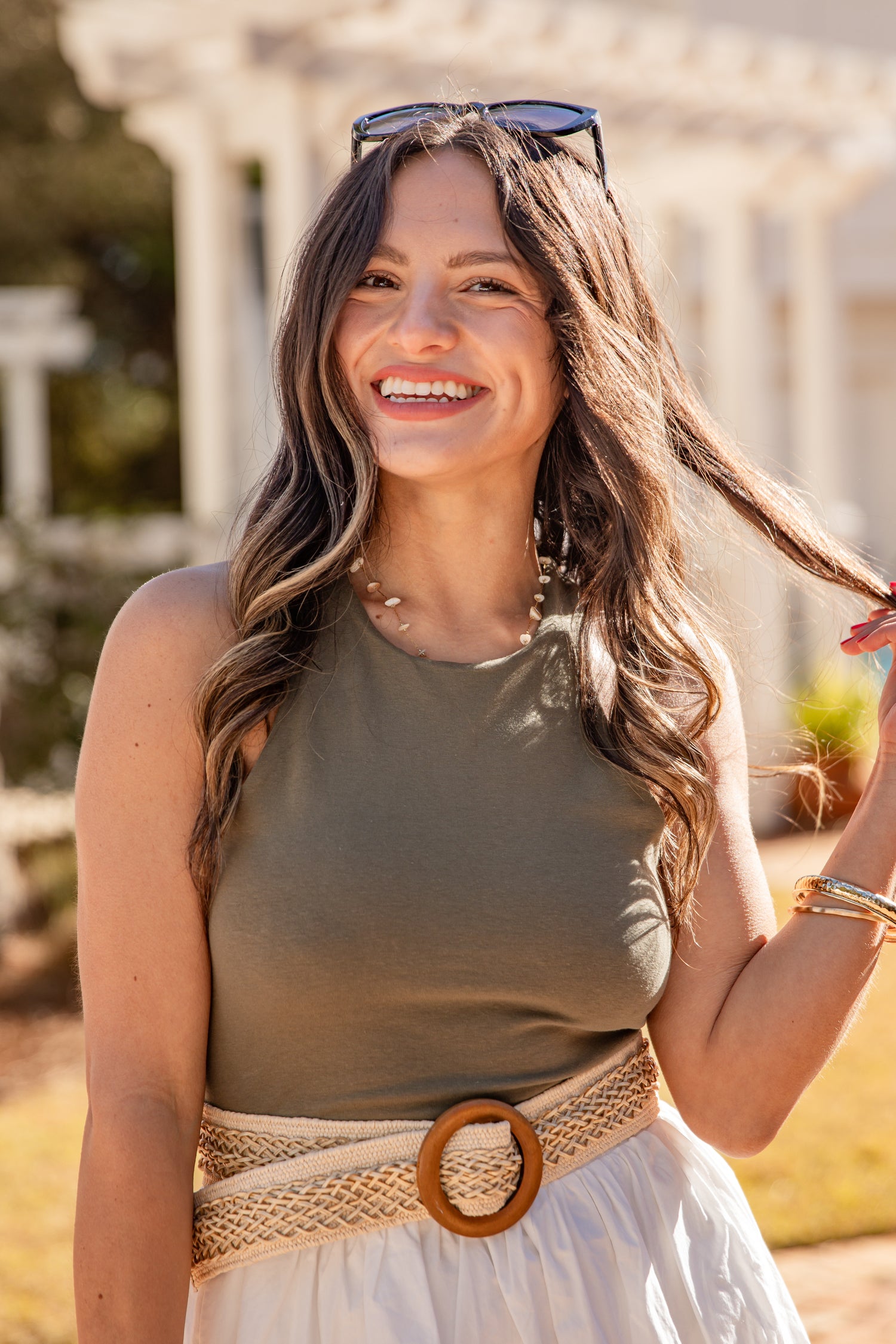 Woman with long hair smiling outdoors, wearing a green top and white skirt.