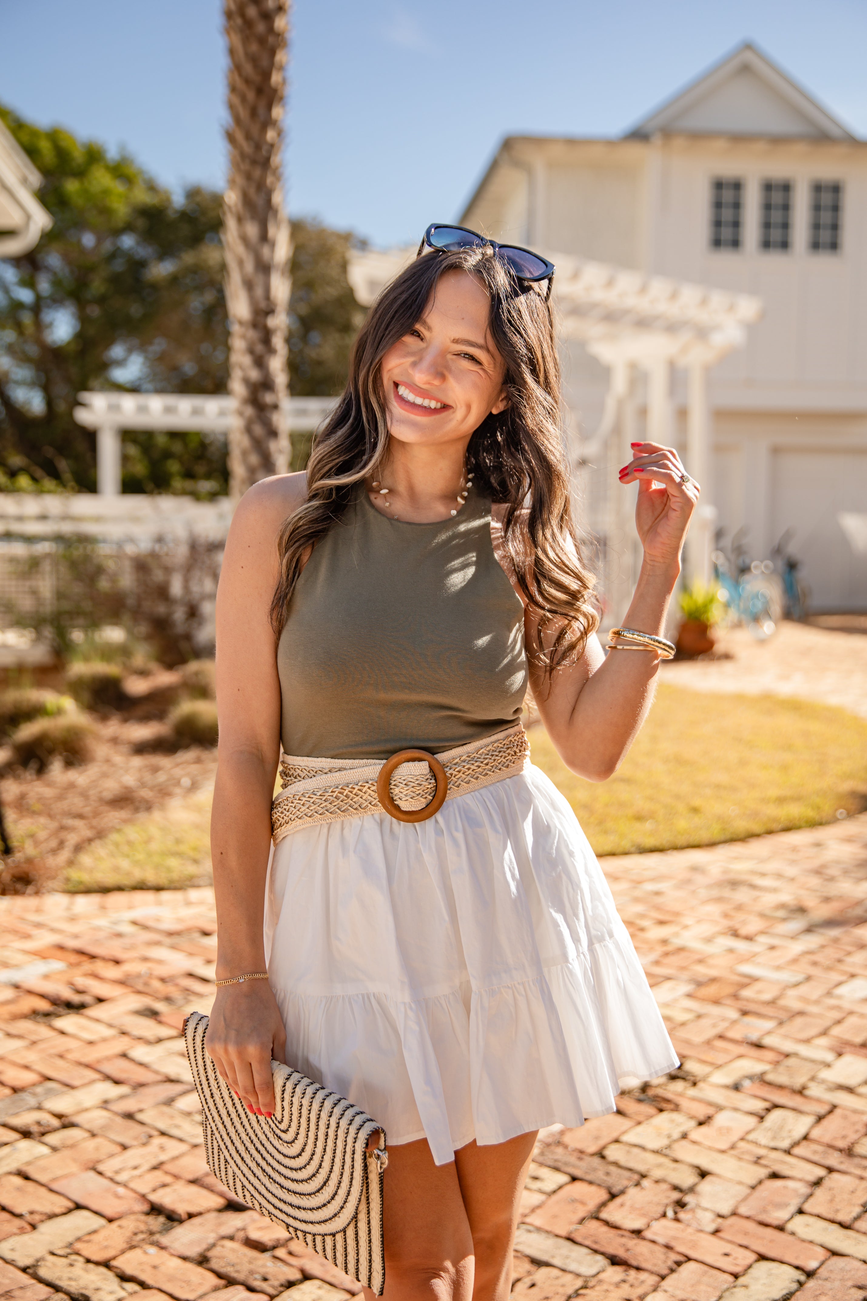 Woman in a green top and white skirt standing on a brick path with palm trees and a building in the background.