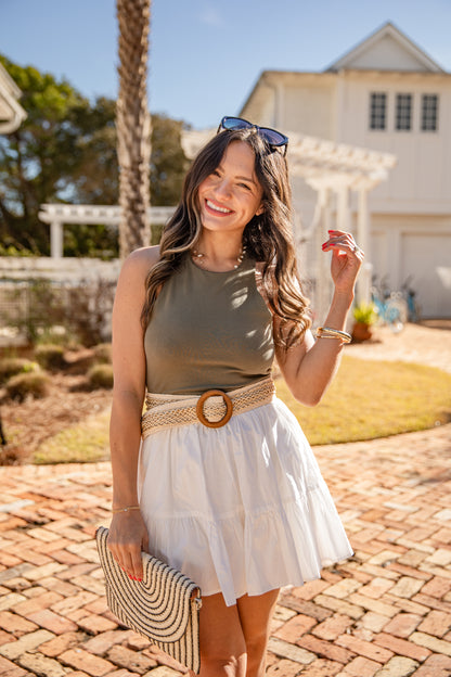 Woman in a green top and white skirt standing on a brick path with palm trees and a building in the background.