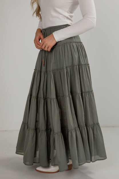 Woman wearing a long, tiered green skirt with a white top on a plain background