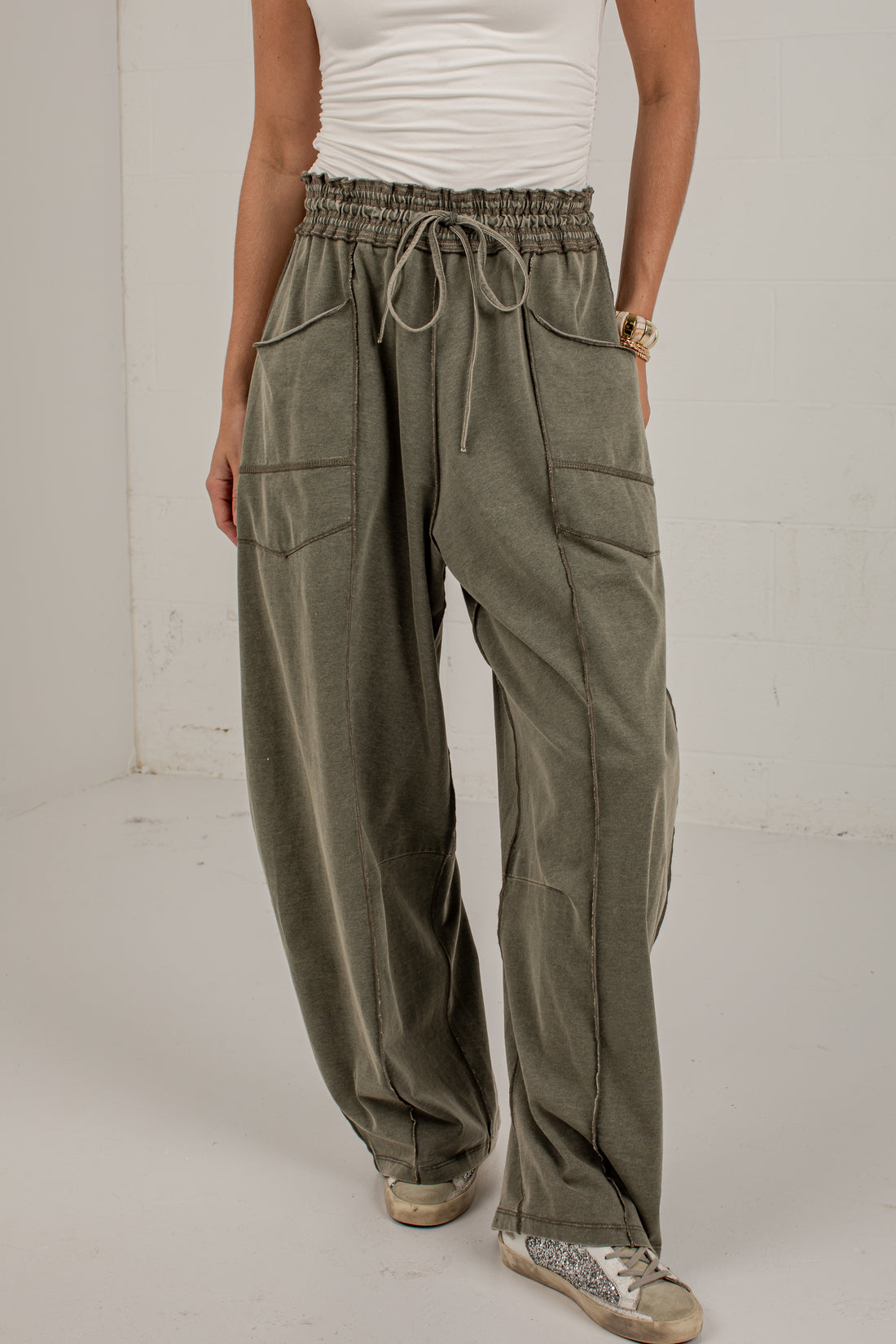 A person wearing loose, wide-leg olive green drawstring pants with large front patch pockets, paired with a white top and beige sneakers, standing against a plain light wall.