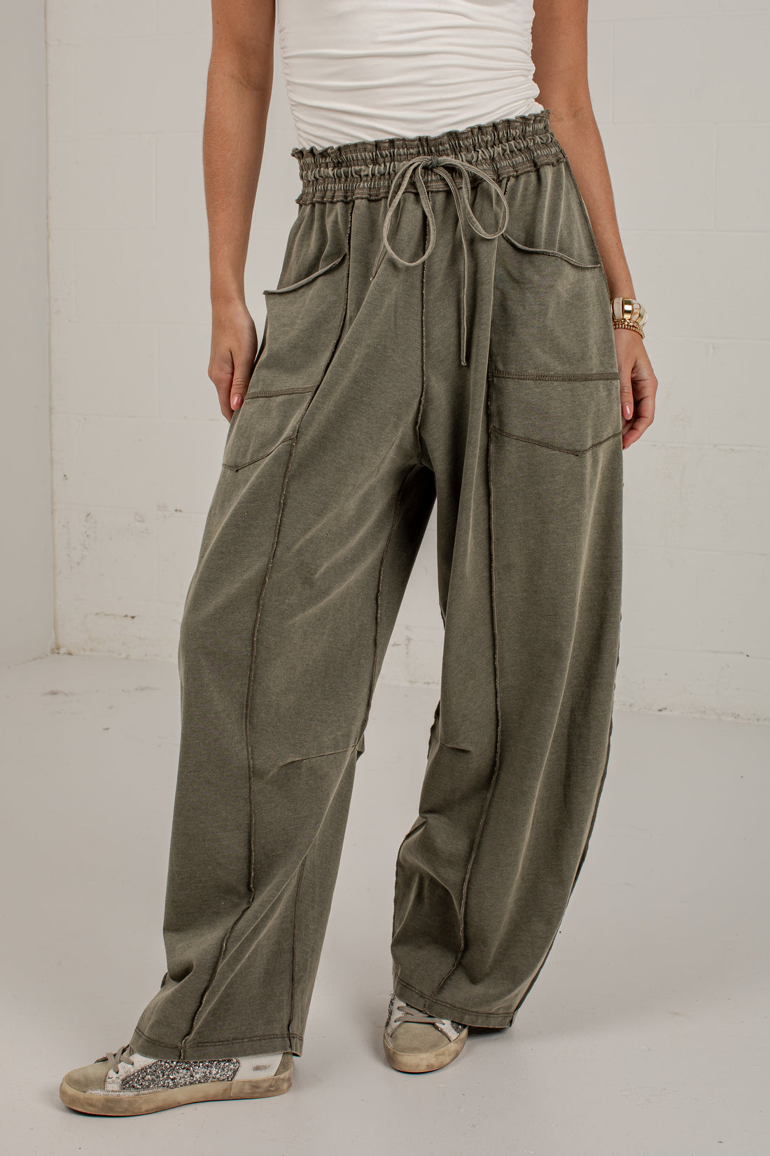 A person wearing loose, wide-leg olive green drawstring pants with large front patch pockets, paired with a white top and beige sneakers, standing against a plain light wall.