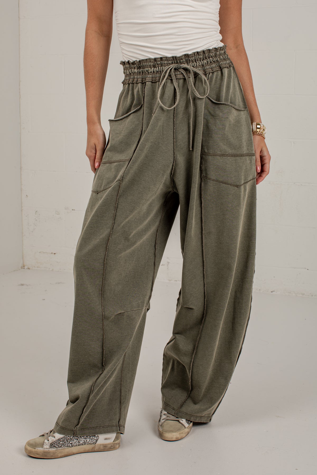 A person wearing loose, wide-leg olive green drawstring pants with large front patch pockets, paired with a white top and beige sneakers, standing against a plain light wall.