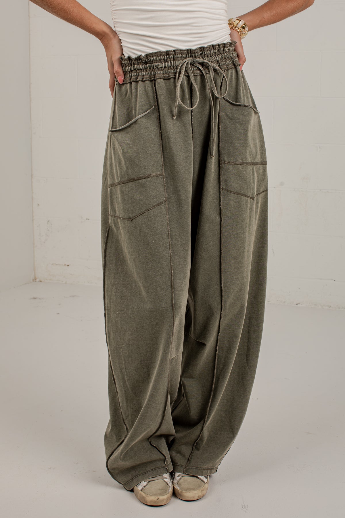 A person wearing very loose, wide-leg olive green drawstring pants with large front patch pockets, paired with a white top and beige shoes, standing against a plain light gray wall.