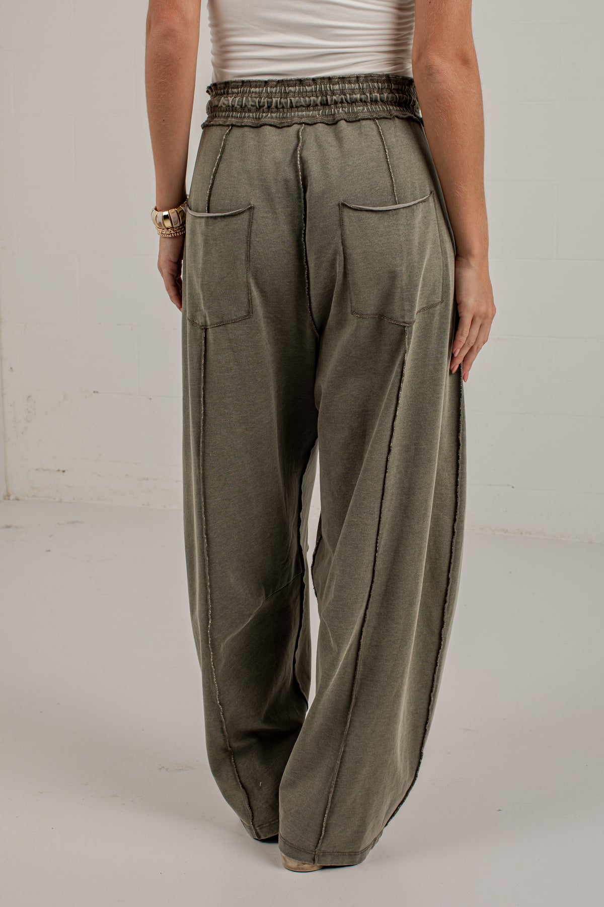 A person wearing loose, wide-leg olive green drawstring pants with large front patch pockets, paired with a white top and beige sneakers, standing against a plain light wall.
