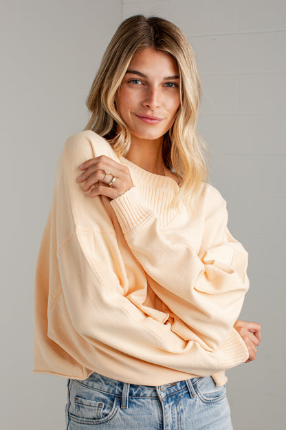 A woman wearing a cream-colored oversized knit sweater and light blue jeans, posing against a light gray wall.