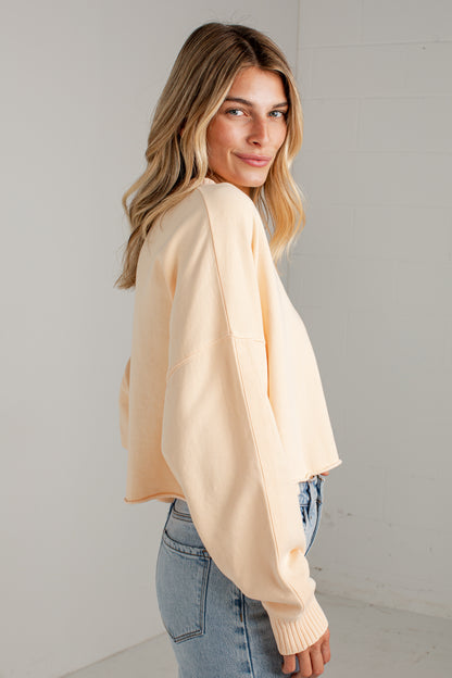 A woman wearing a light peach-colored cropped knit sweater and light blue jeans, photographed from the back-three-quarter view against a plain white wall.
