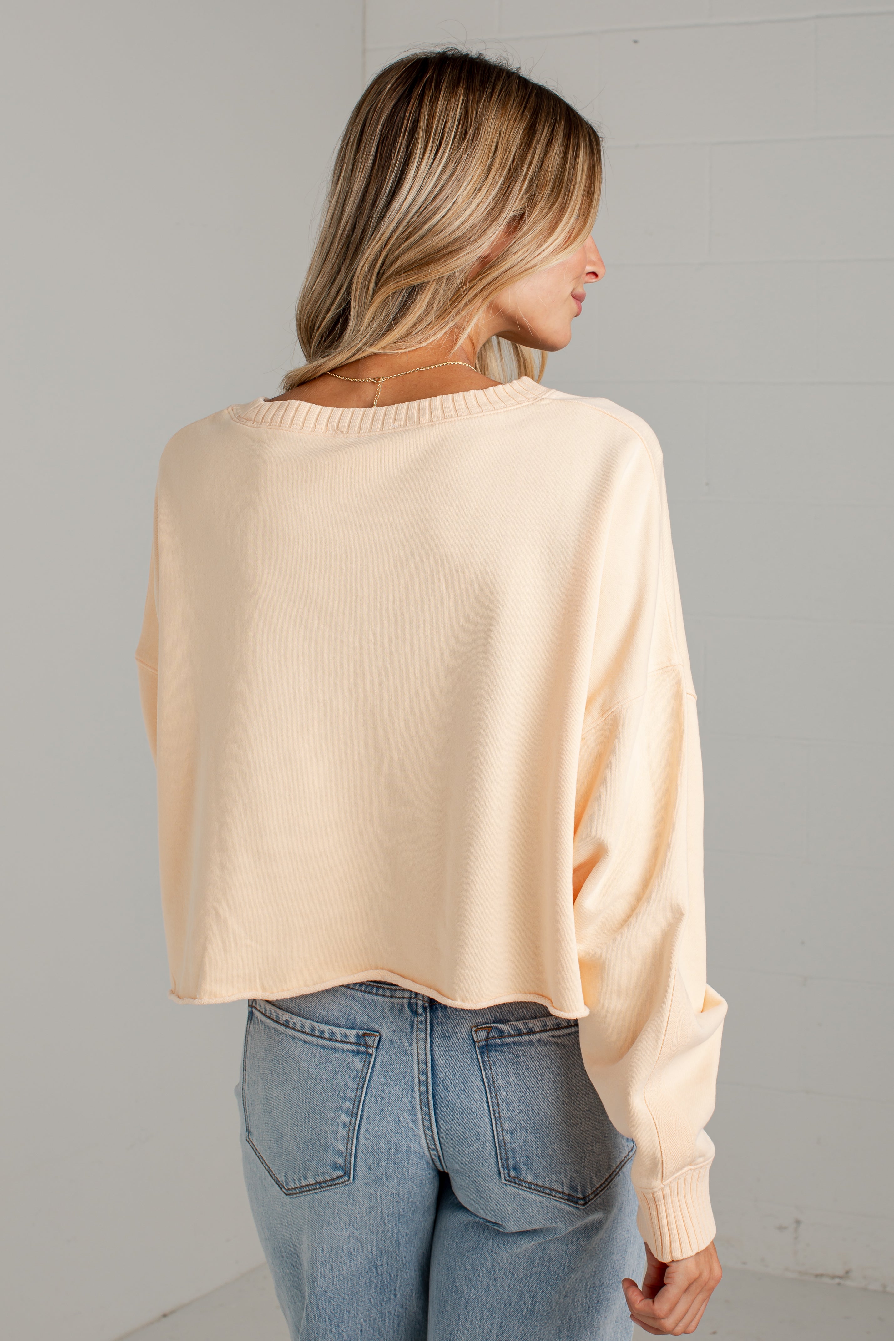 A woman shown from the back wearing a light peach-beige long-sleeve knit sweater and blue jeans against a light gray wall.