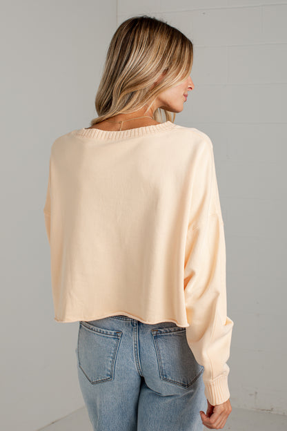 A woman shown from the back wearing a light peach-beige long-sleeve knit sweater and blue jeans against a light gray wall.