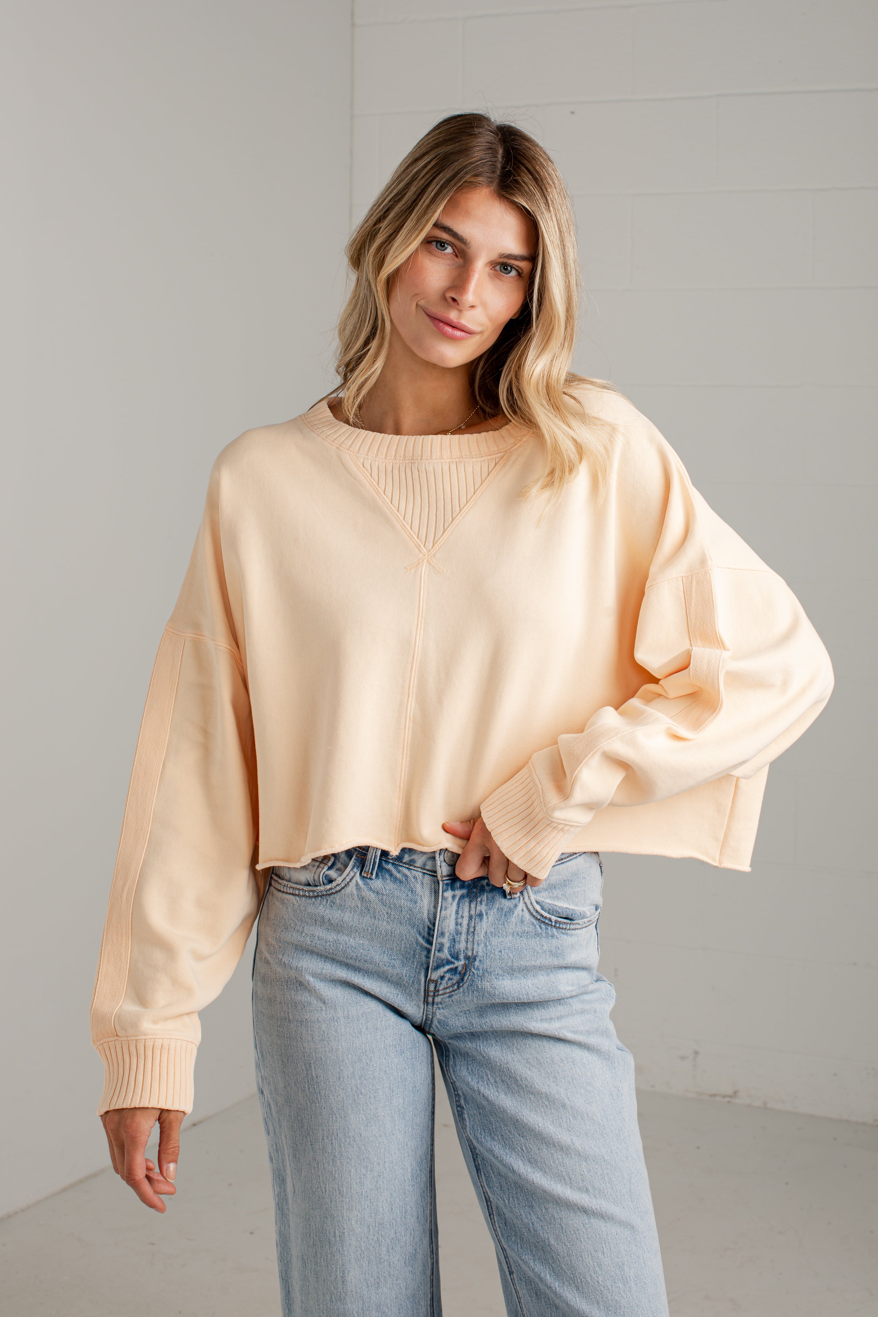 A woman wearing a light peach-colored knit sweater with balloon sleeves and light blue jeans, standing against a white paneled wall.