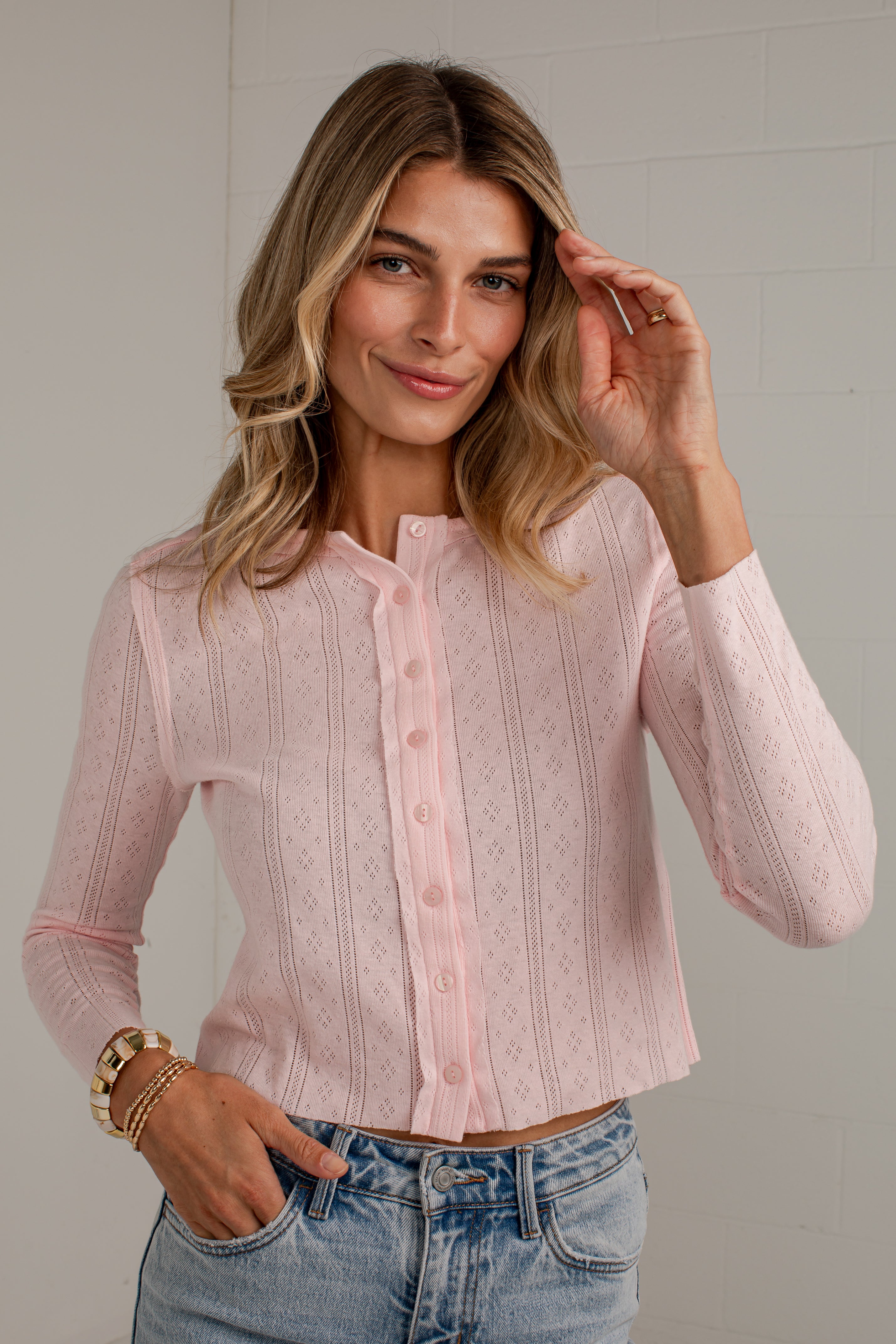 A woman wearing a light pink long-sleeve button-front knit top with a stand collar, paired with light-wash blue jeans, standing against a light gray wall.