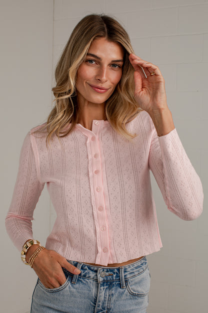 A woman wearing a light pink long-sleeve button-front knit top with a stand collar, paired with light-wash blue jeans, standing against a light gray wall.