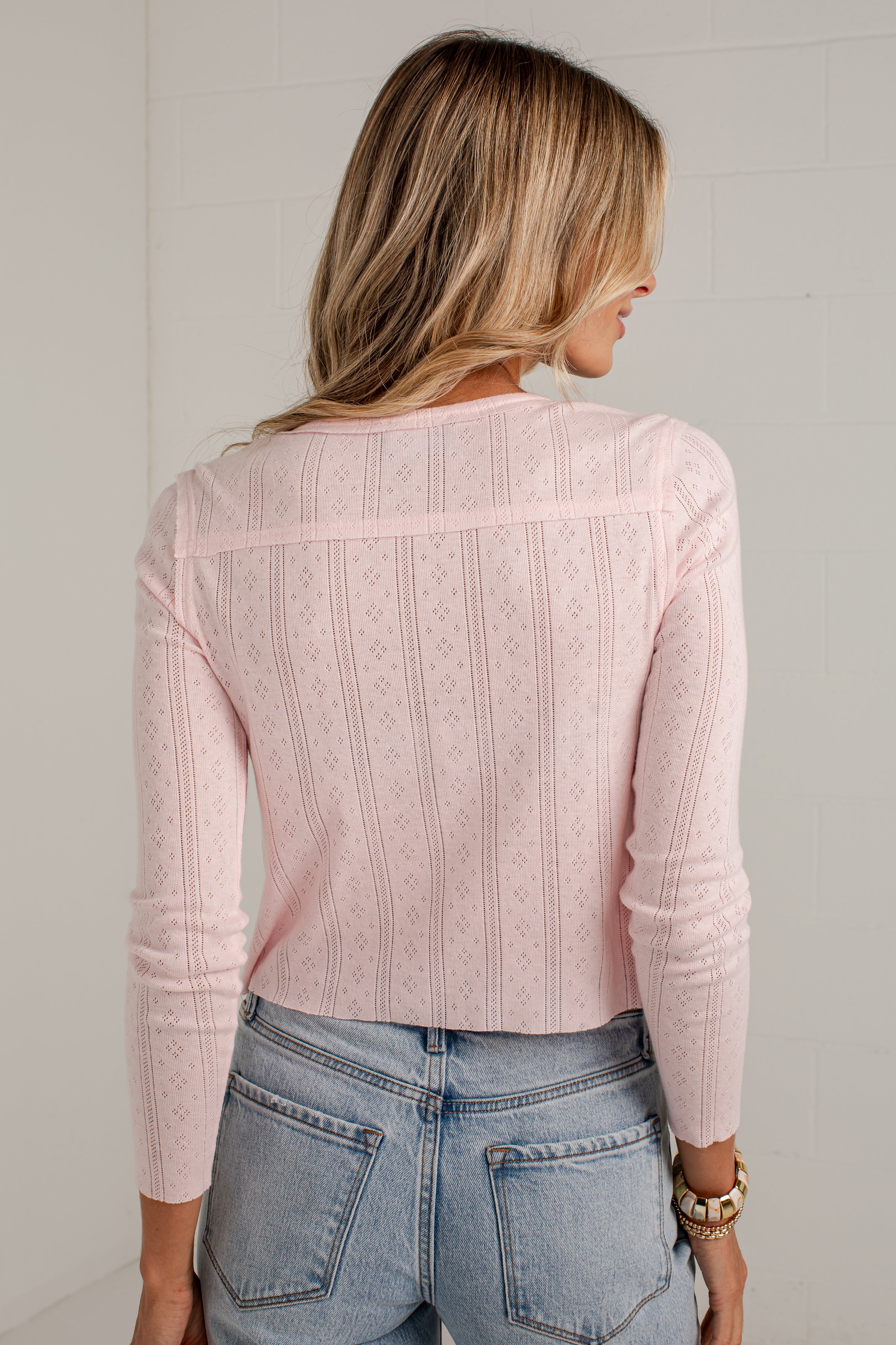 A woman viewed from behind wearing a light pink long-sleeve knit sweater with textured vertical panels and light-wash blue jeans, standing against a white paneled wall.