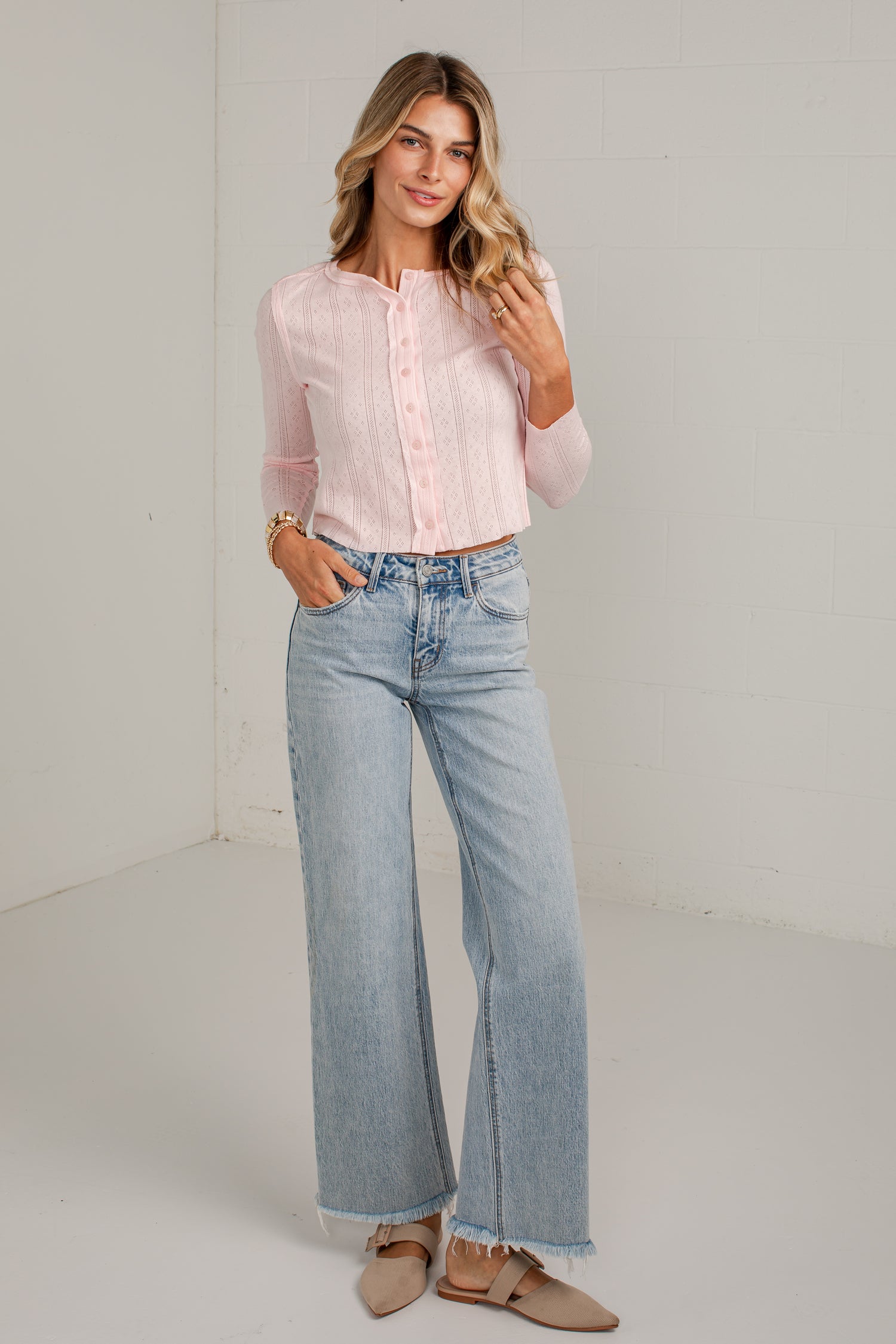 A woman modeling a light pink long-sleeve button-up blouse and light-wash wide-leg jeans, standing in a bright white studio corner.