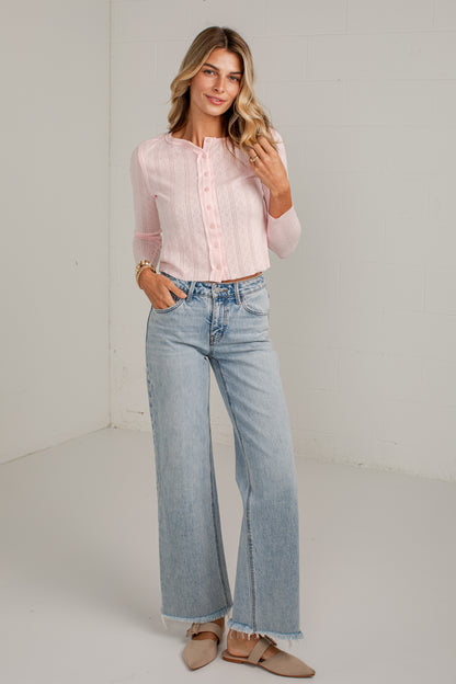 A woman modeling a light pink long-sleeve button-up blouse and light-wash wide-leg jeans, standing in a bright white studio corner.