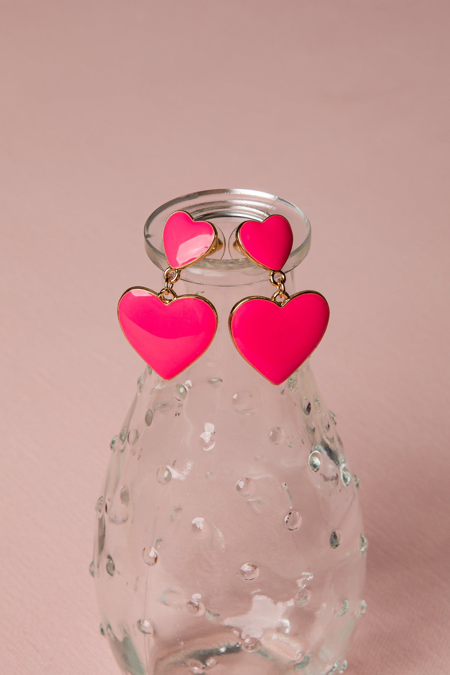 Pink heart-shaped earrings on a clear glass bottle with a pink background