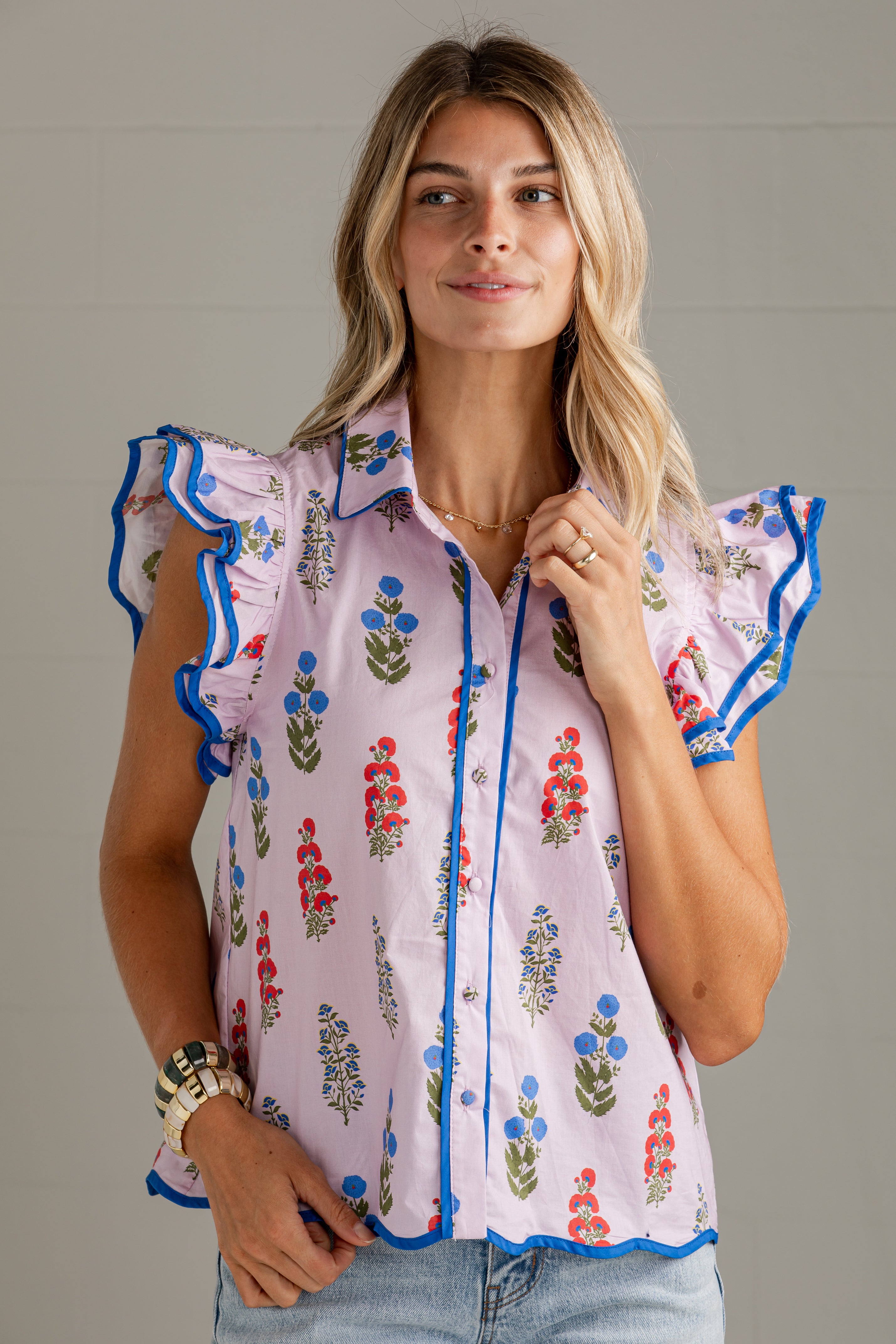 A woman wearing a light pink sleeveless blouse with blue ruffle trim and a colorful floral print, paired with light blue jeans, against a neutral gray background.
