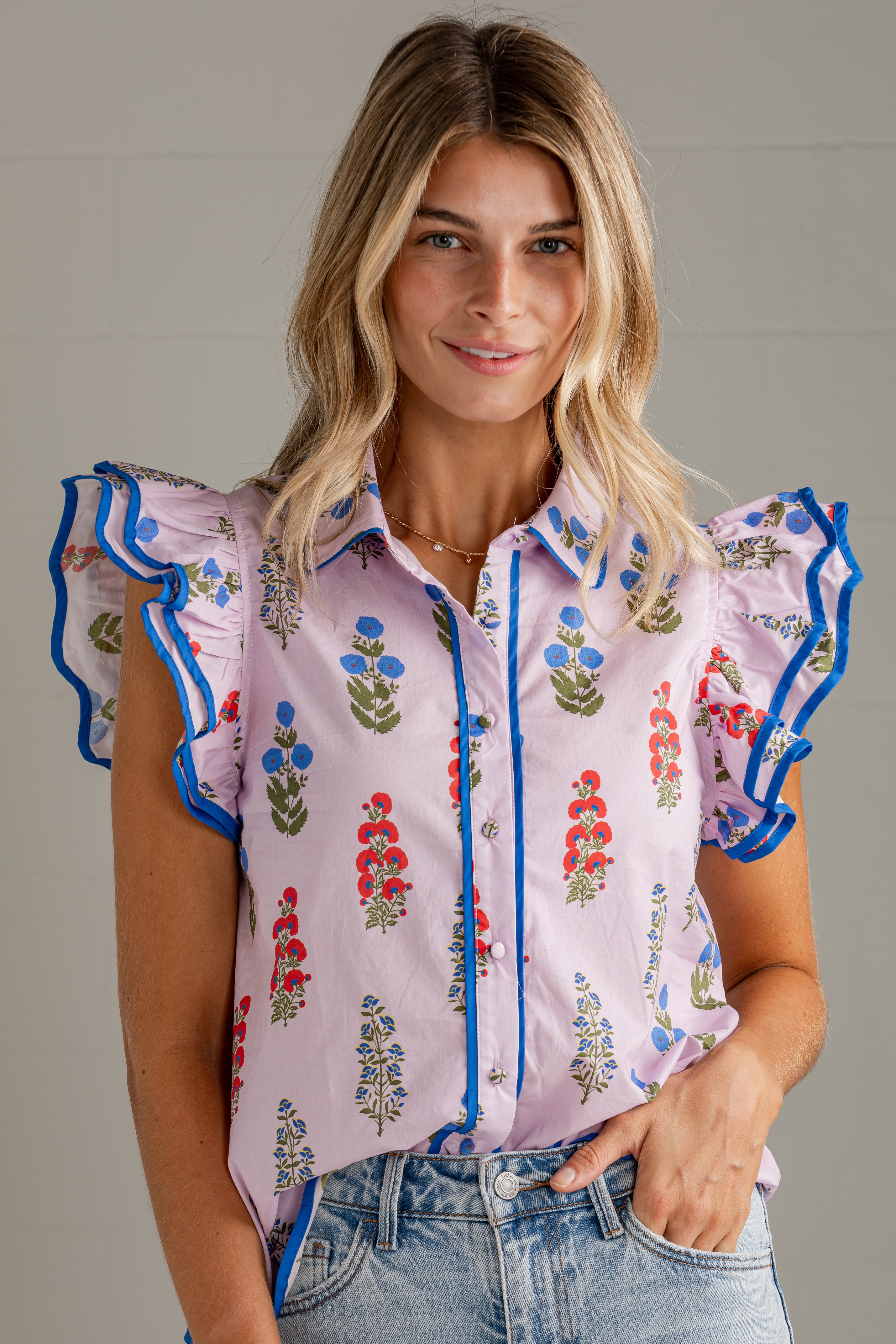 A woman wearing a light pink ruffled blouse with blue trim and a colorful floral print, paired with light-wash denim jeans, against a neutral gray background.