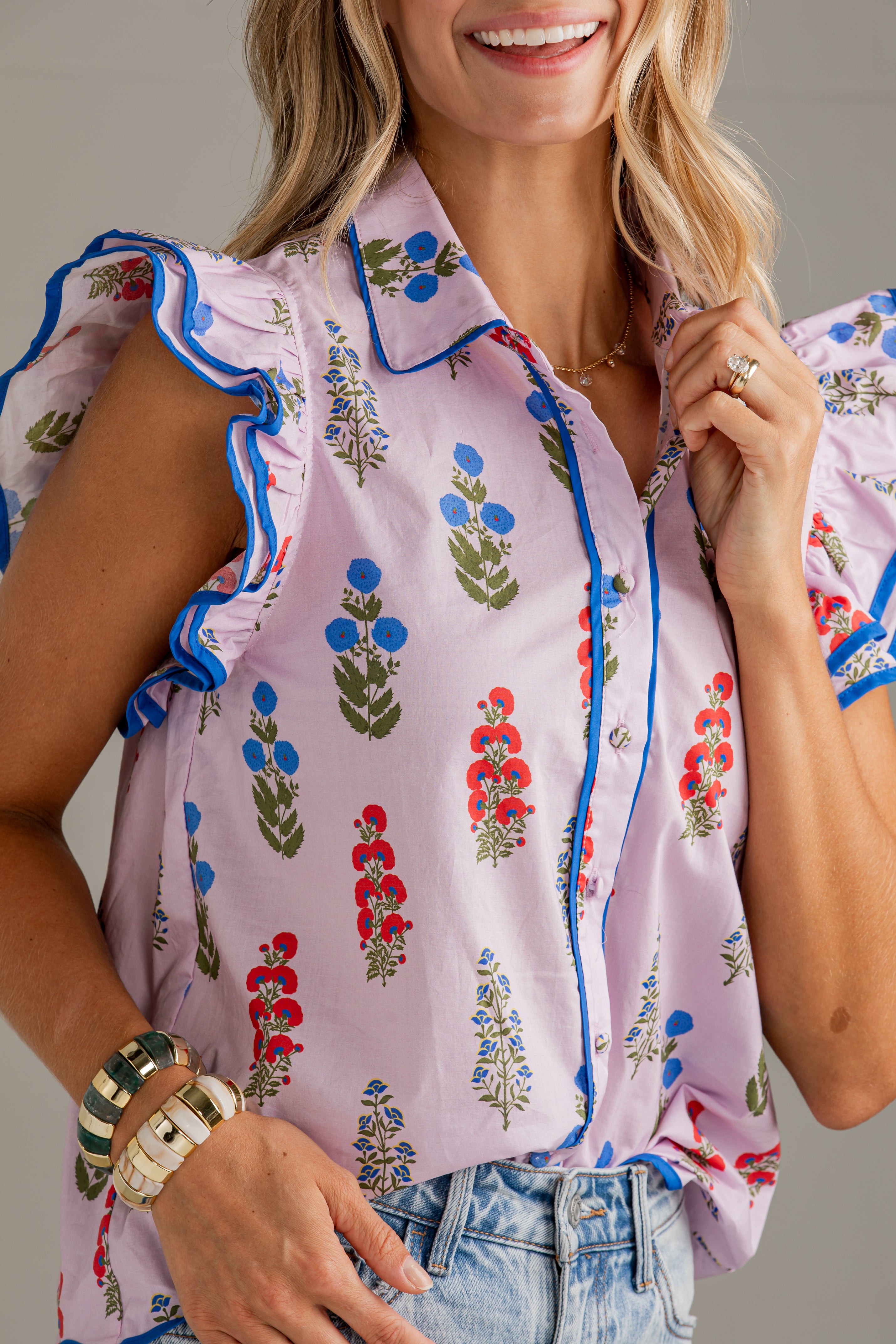 A close-up of a woman wearing a light pink sleeveless blouse with blue and red floral prints and ruffled trim, paired with light-wash denim jeans against a neutral gray background.