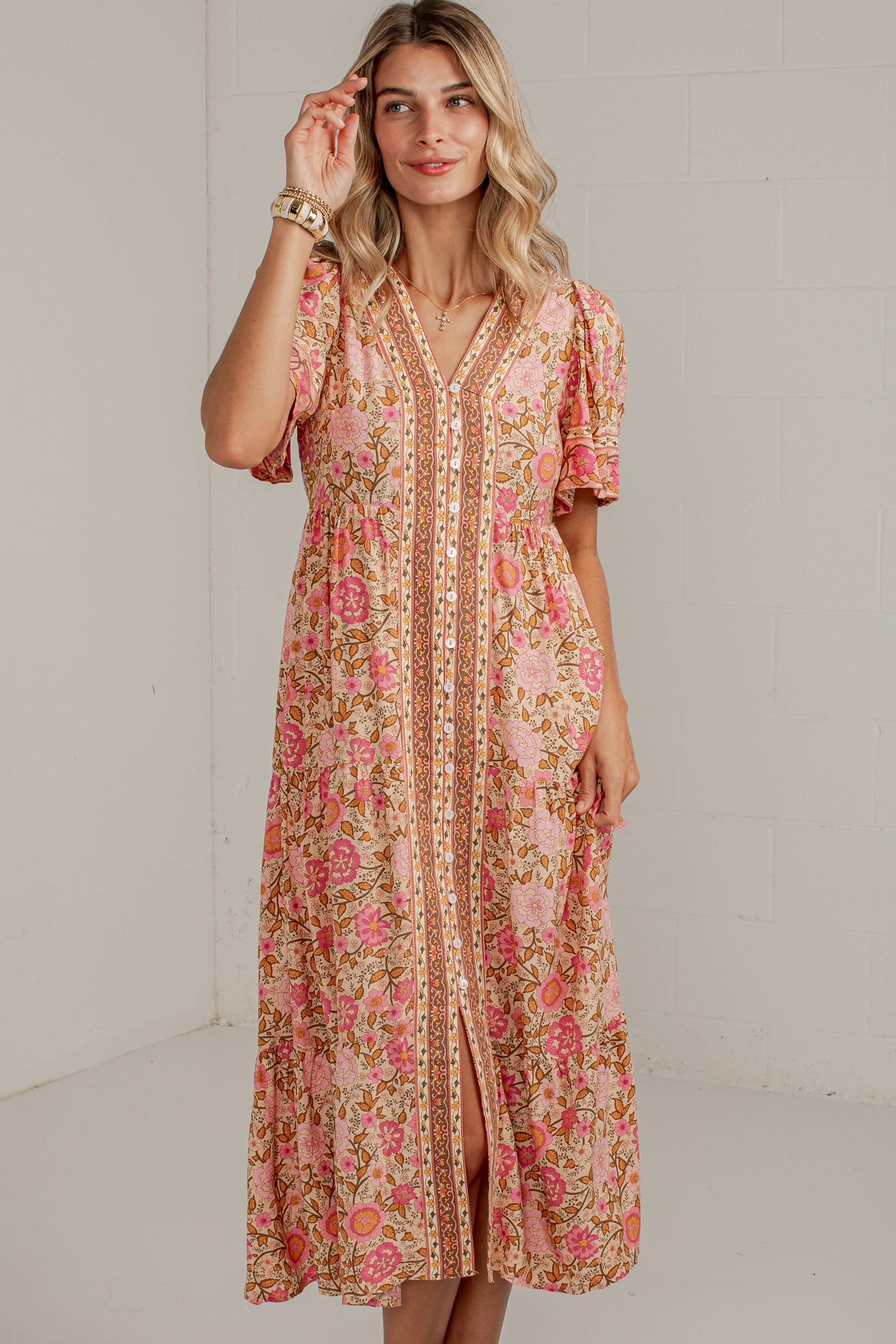 A woman modeling a peach and pink floral midi dress with gold embroidery down the front, standing in a white studio setting.