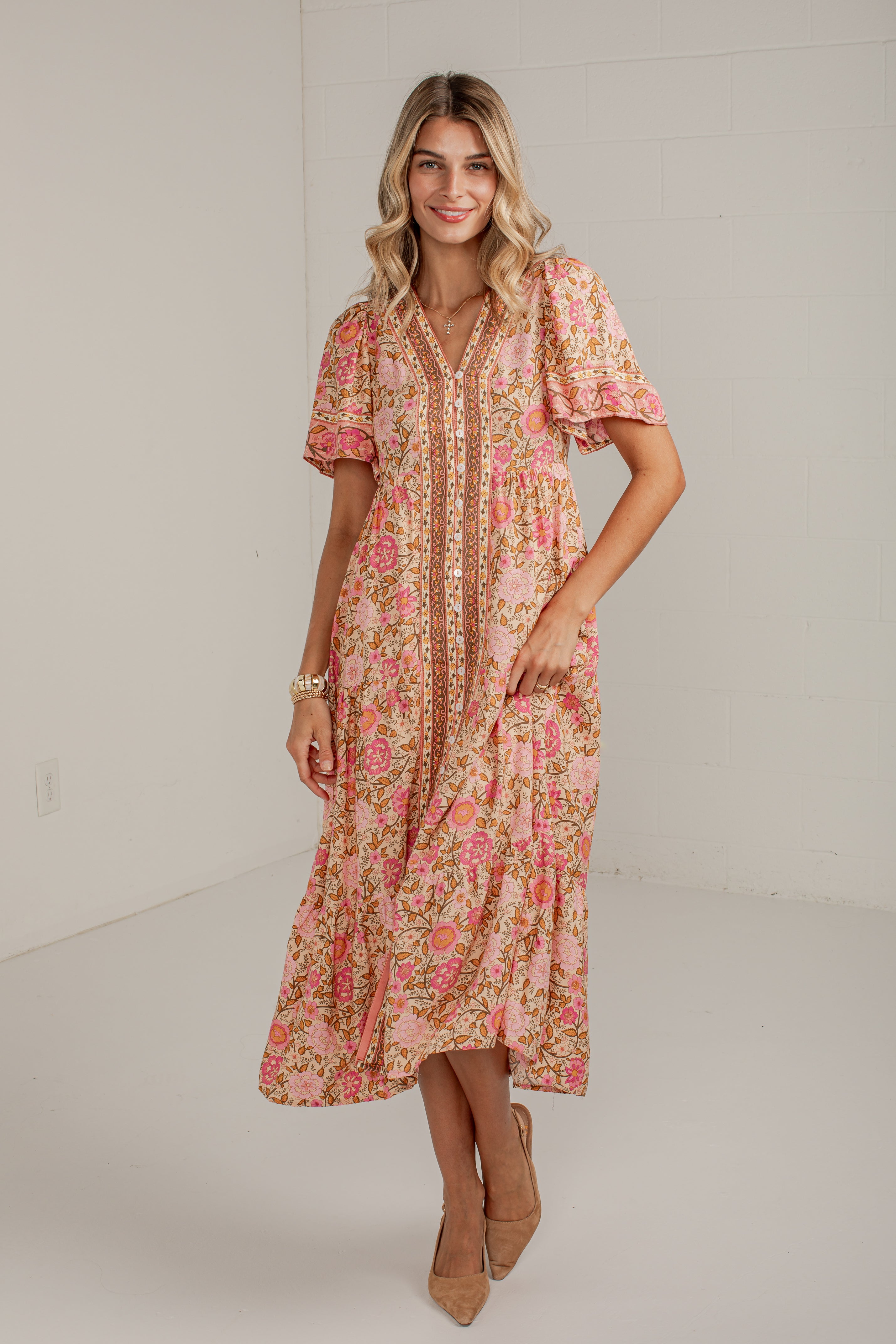 A woman modeling a short-sleeve, tiered midi dress with a pink and orange floral print on a beige background, styled with tan heels in a bright studio setting.