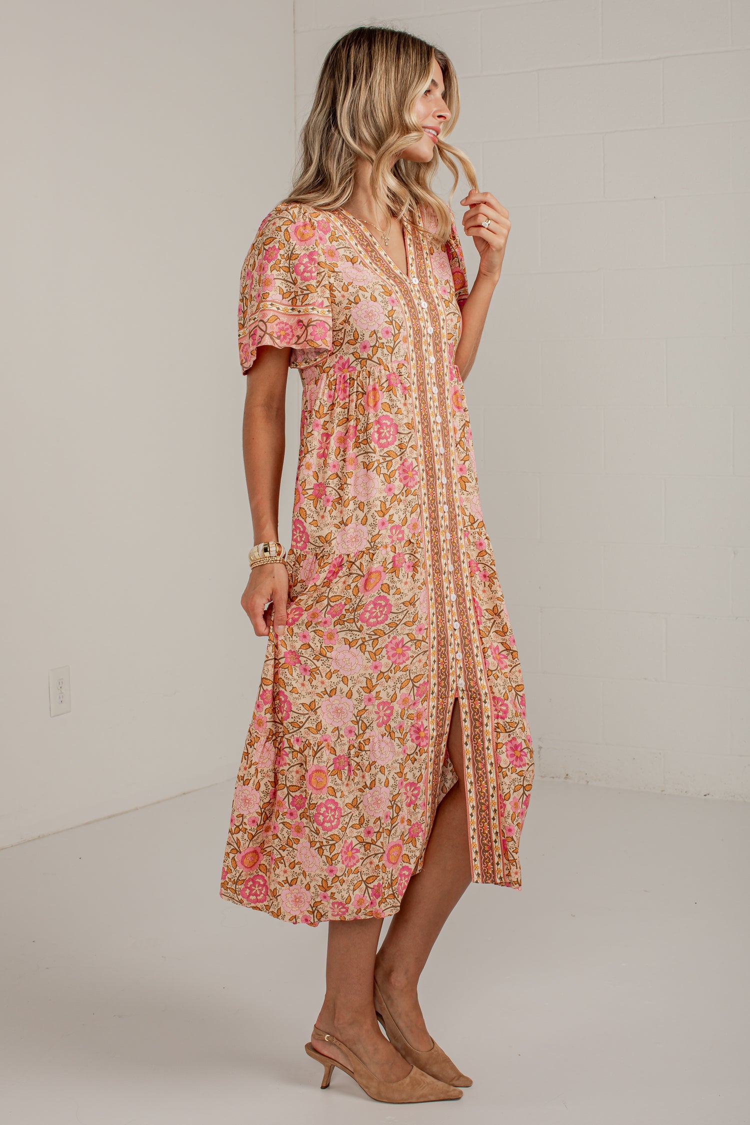 A woman modeling a peach and pink floral midi dress with a split front and beige trim, standing in a bright white studio.