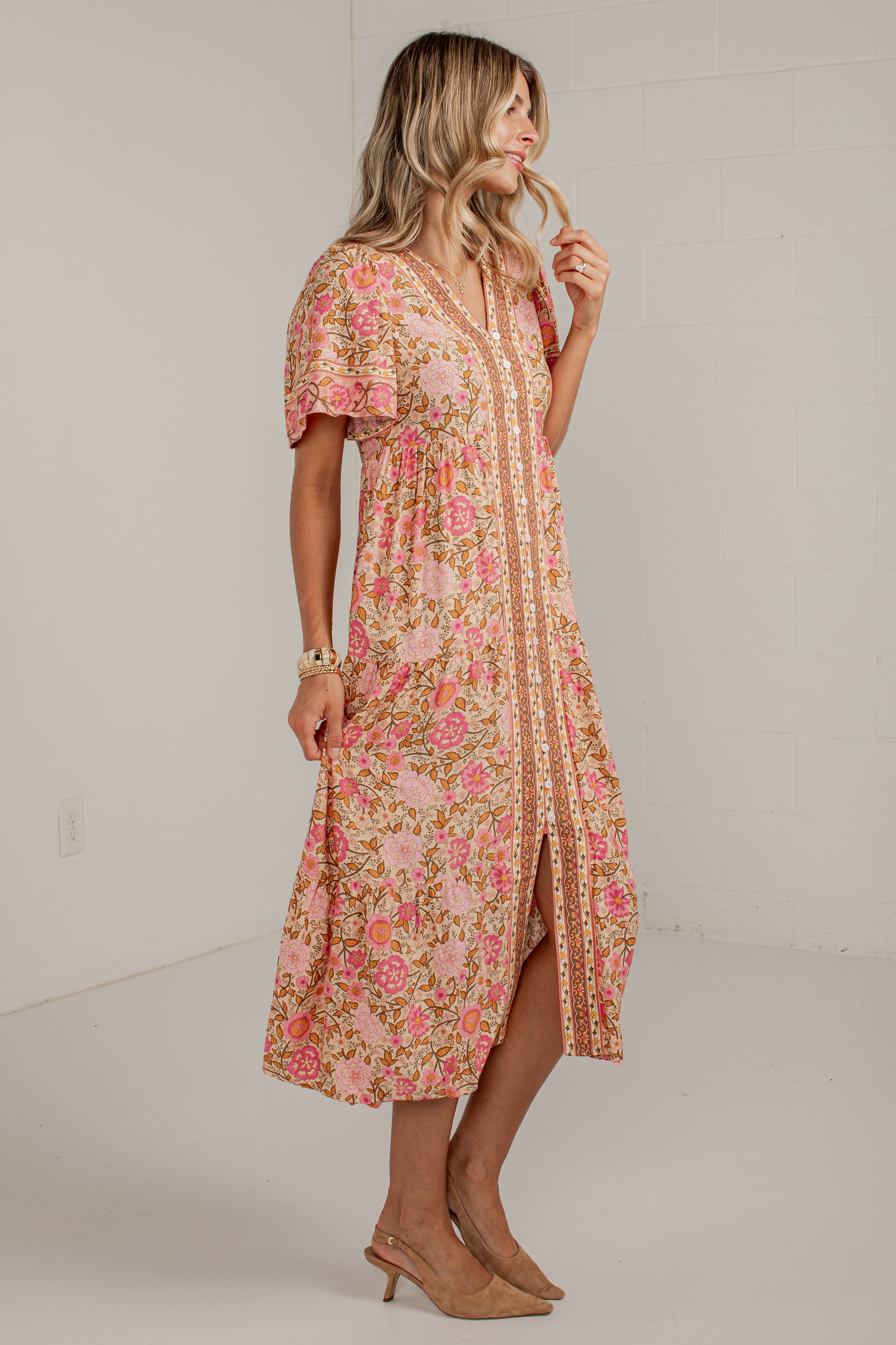 A woman modeling a peach and pink floral midi dress with a split front and beige trim, standing in a bright white studio.