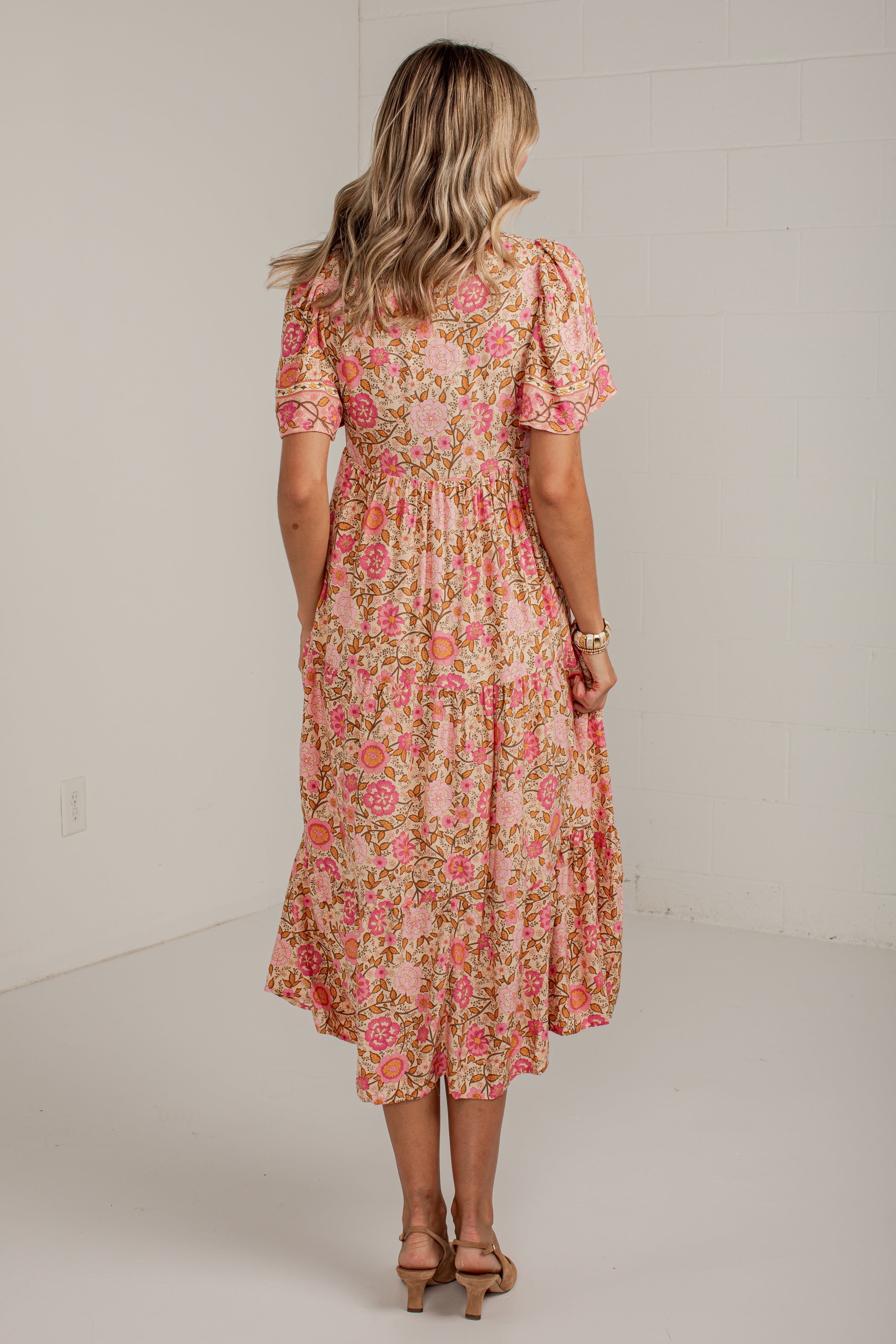 A woman viewed from behind wearing a short-sleeve midi dress with a pink and green floral pattern, standing in a bright studio with a white wall.