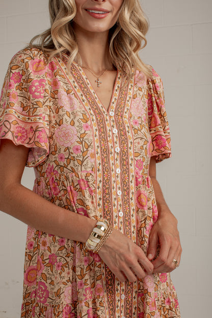 A woman wearing a short-sleeve bohemian-style dress with a pink, orange, and beige floral print, featuring a button-down front and ruffled sleeves.