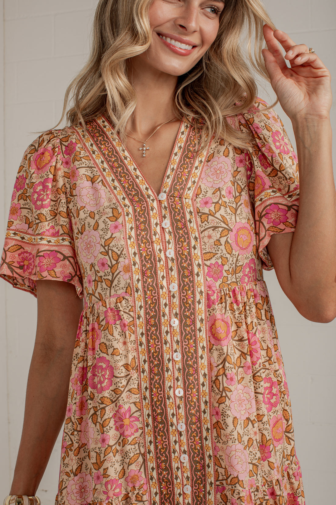 A woman wearing a bohemian-style short-sleeve dress with a pink, orange, and beige floral print and vertical patterned borders, shown in a close-up shot against a light gray wall.