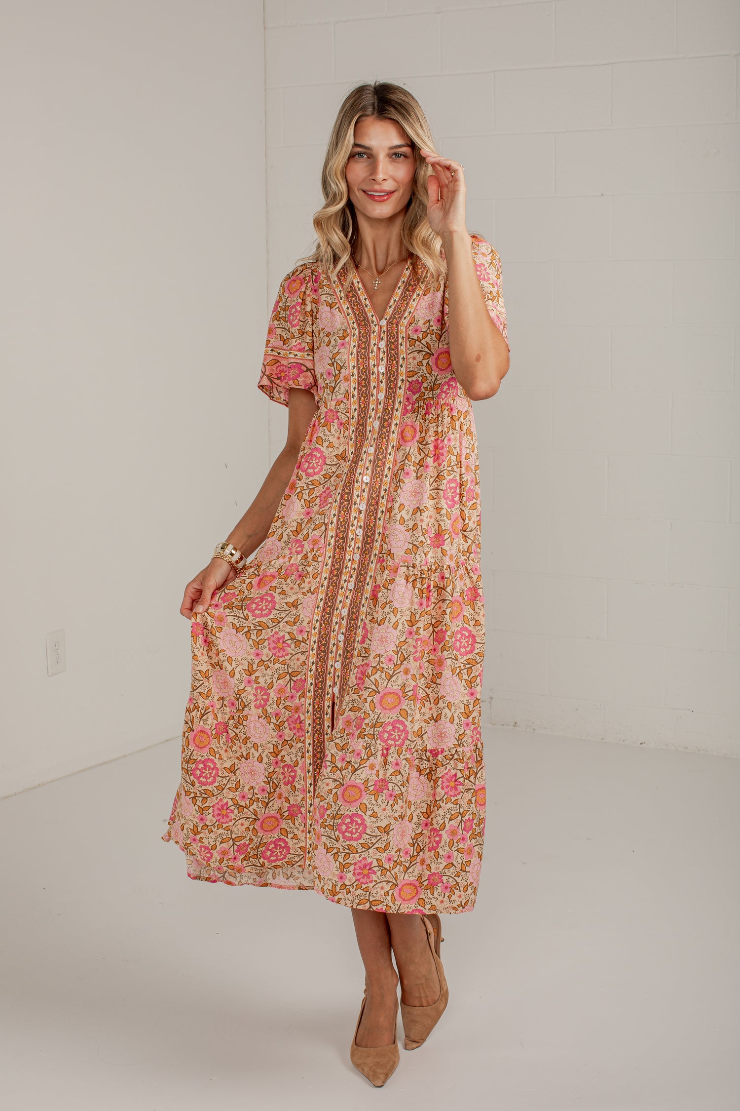A woman modeling a short-sleeve midi dress with a pink and green floral print and a gold-toned embroidered tie-front detail, standing against a white studio wall.