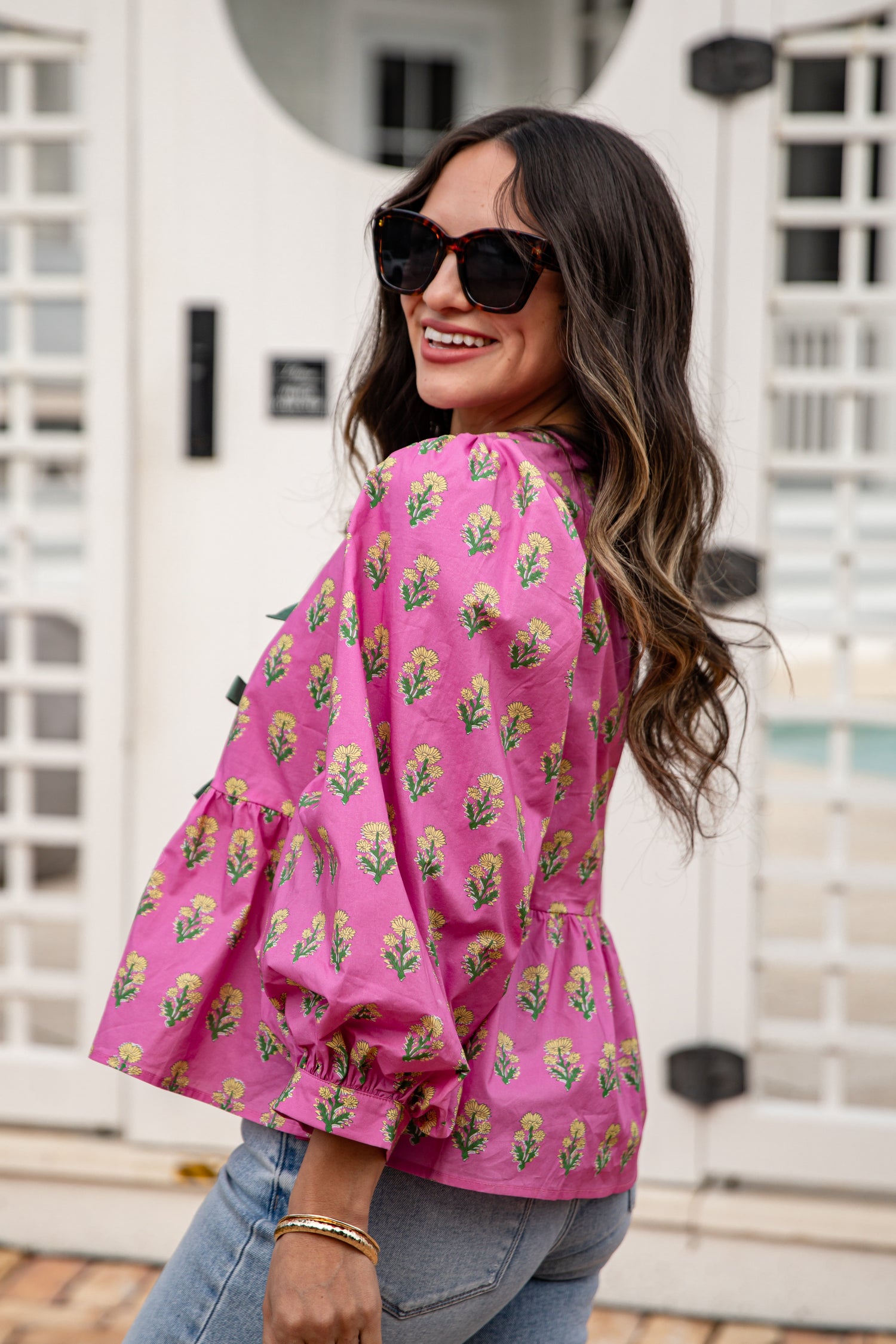 Woman wearing a pink floral blouse with sunglasses, standing in front of a white building.