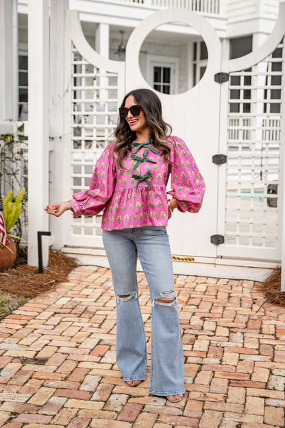 Woman wearing a pink blouse with floral patterns and light blue jeans standing on a brick patio.