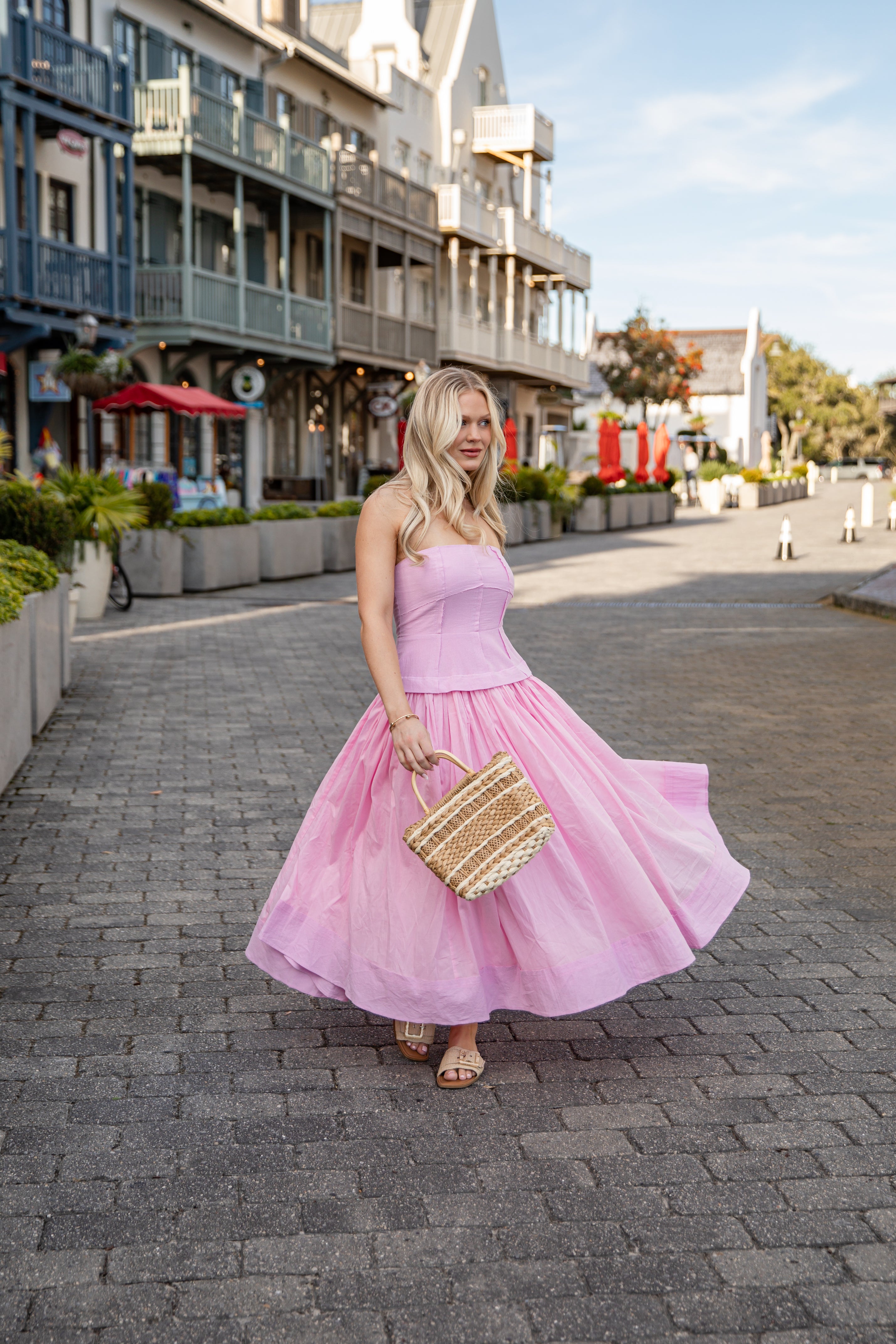 Woman in a pink dress holding a straw bag on a street with buildings and plants in the background