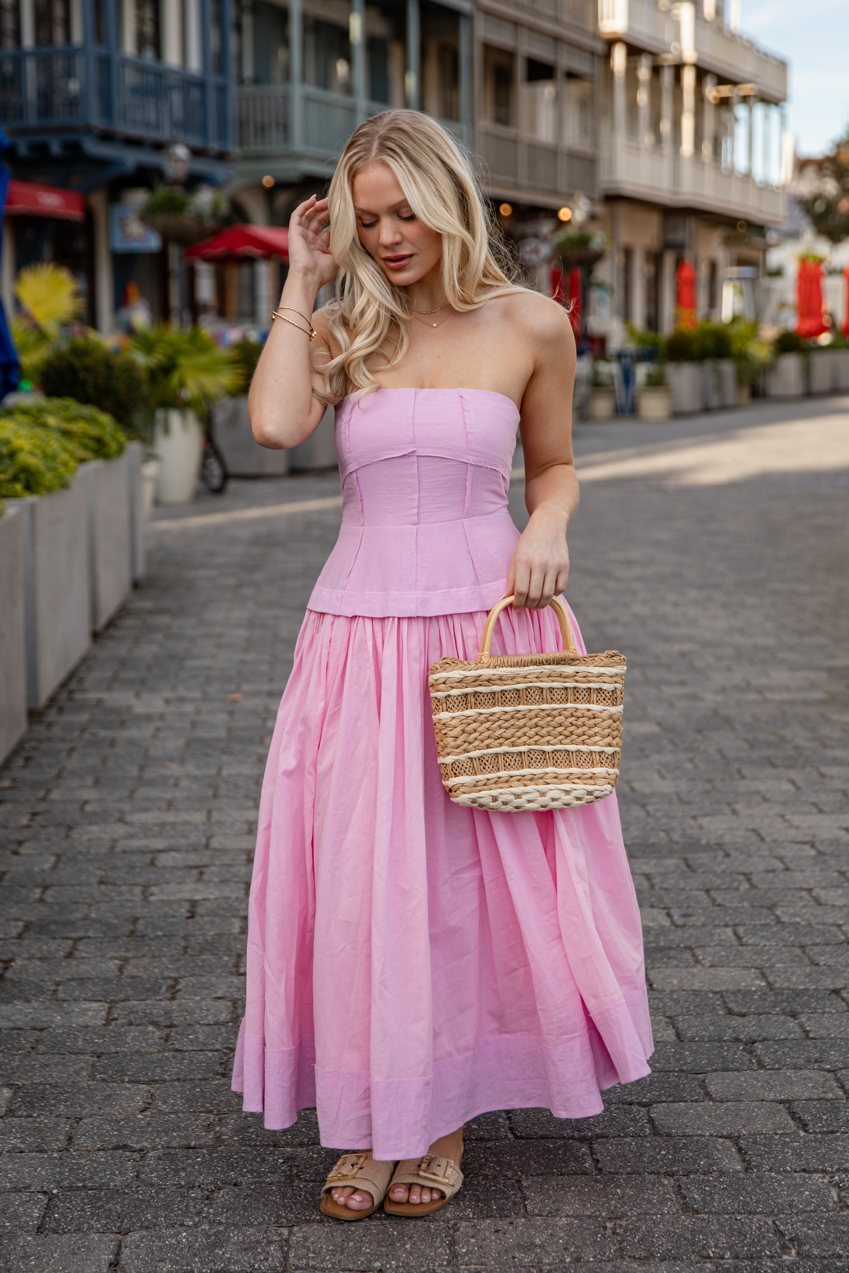 Woman in a pink strapless dress holding a straw bag on a street.