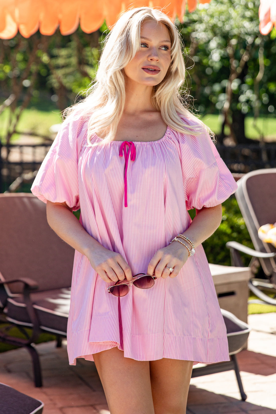 Woman in a pink dress standing outdoors with greenery and furniture in the background