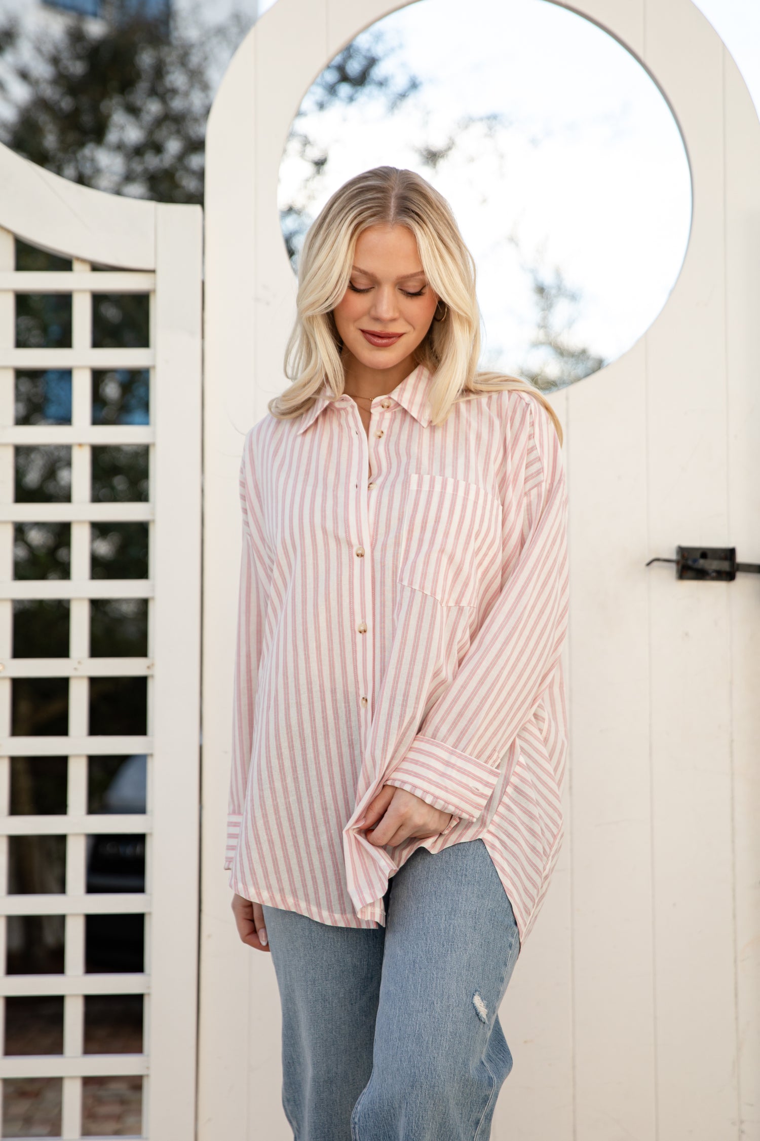 Woman wearing a pink striped shirt and blue jeans standing in front of a white lattice gate.