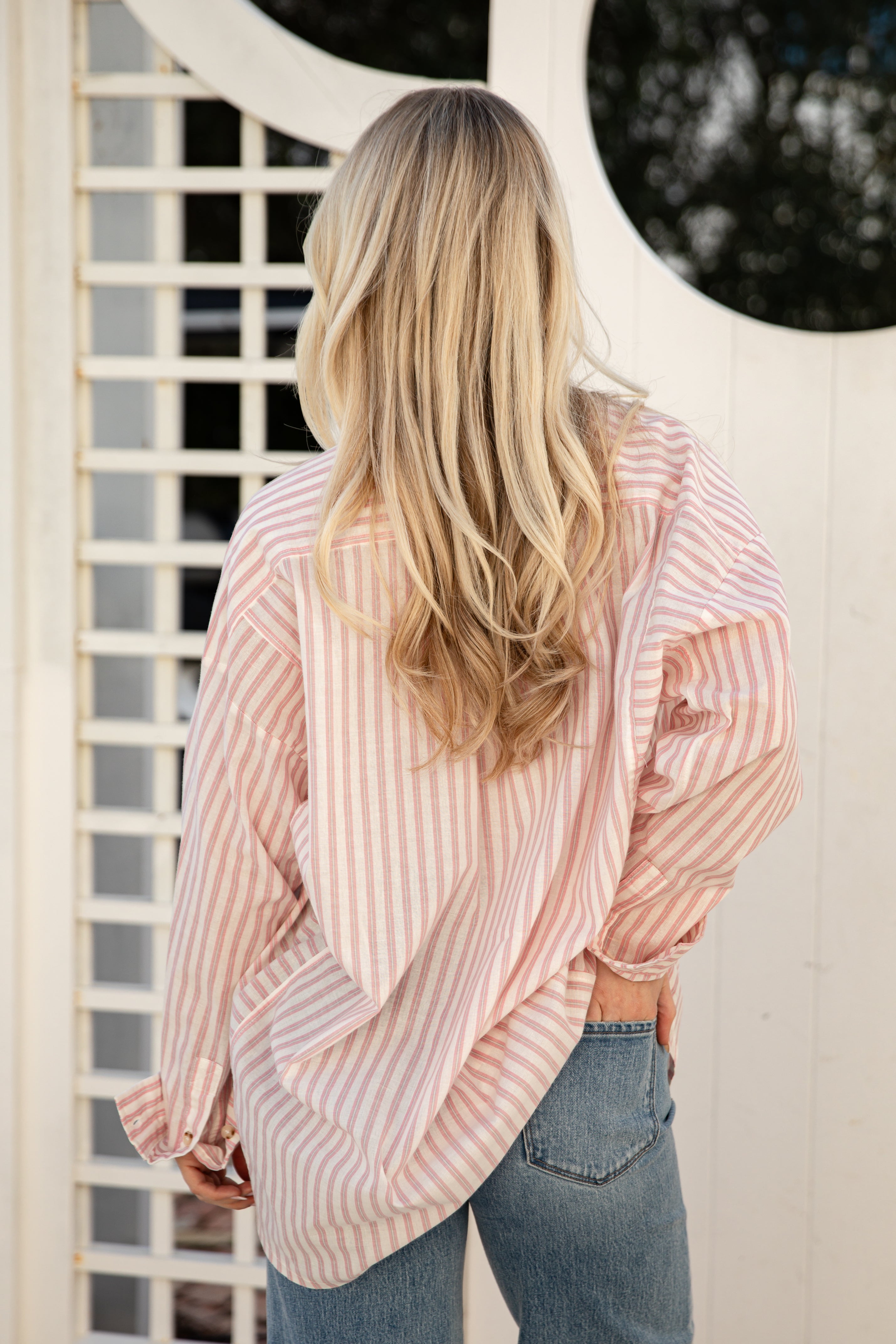 Person wearing a pink and white striped shirt and blue jeans standing in front of a white lattice door.
