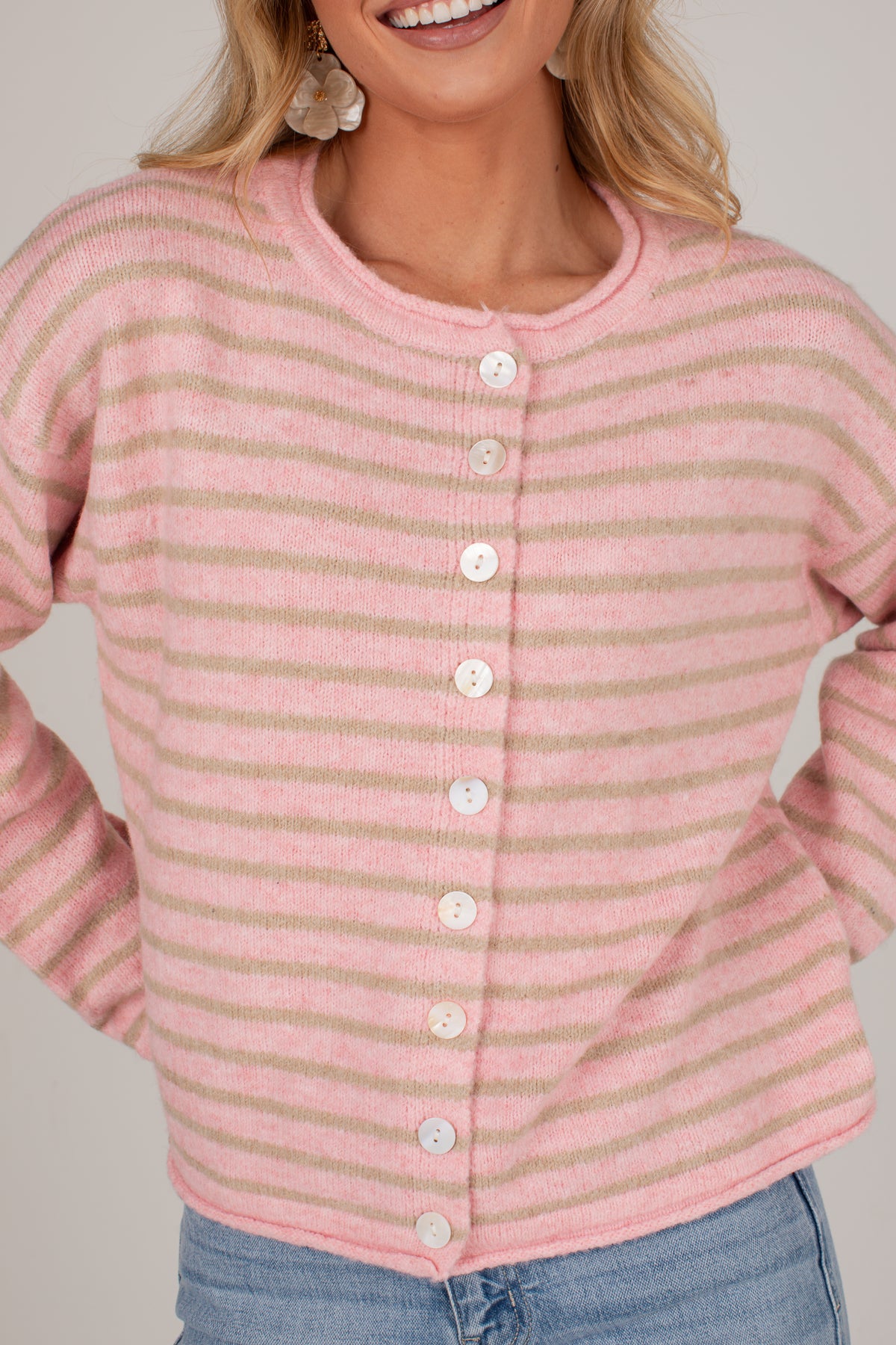 A close-up of a woman wearing a pink and beige striped knit cardigan with white buttons, paired with blue jeans against a light gray background.