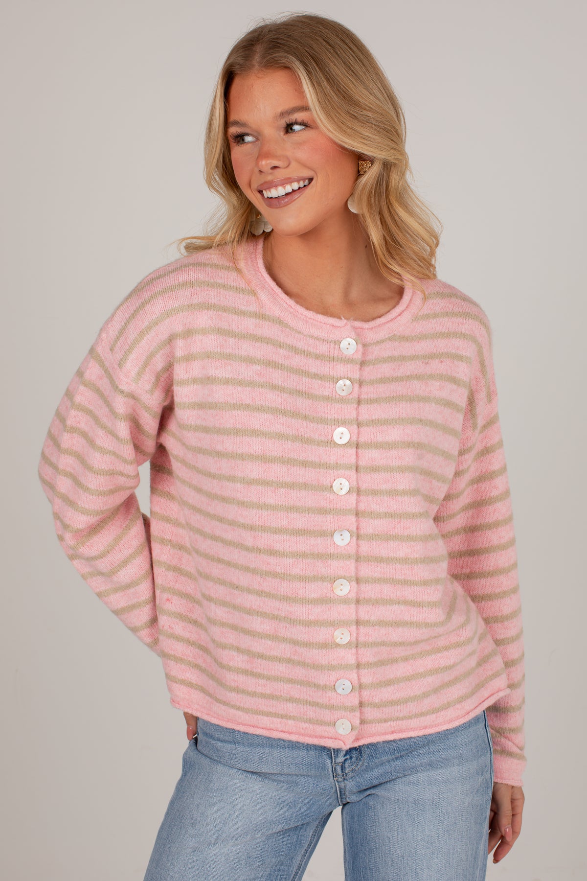A woman wearing a pink and light beige striped knit cardigan with white buttons, paired with blue jeans, against a plain light gray studio background.