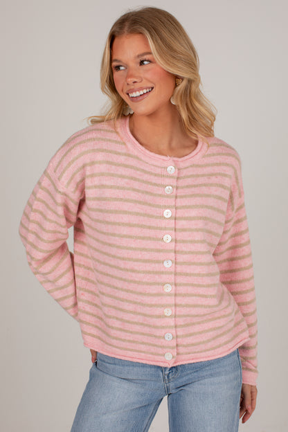 A woman wearing a pink and light beige striped knit cardigan with white buttons, paired with blue jeans, against a plain light gray studio background.
