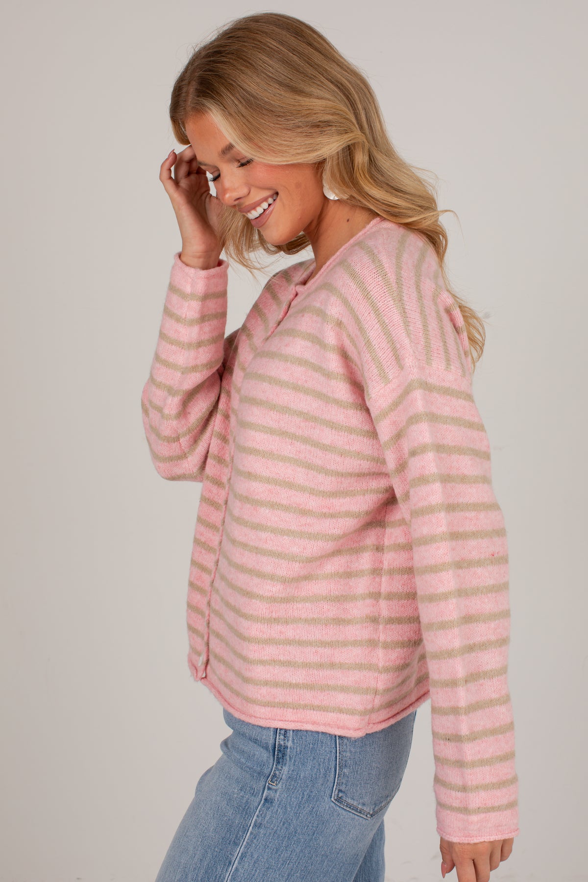 A woman wearing a pink and beige striped knit sweater with blue jeans, shown from a side profile against a light gray studio background.