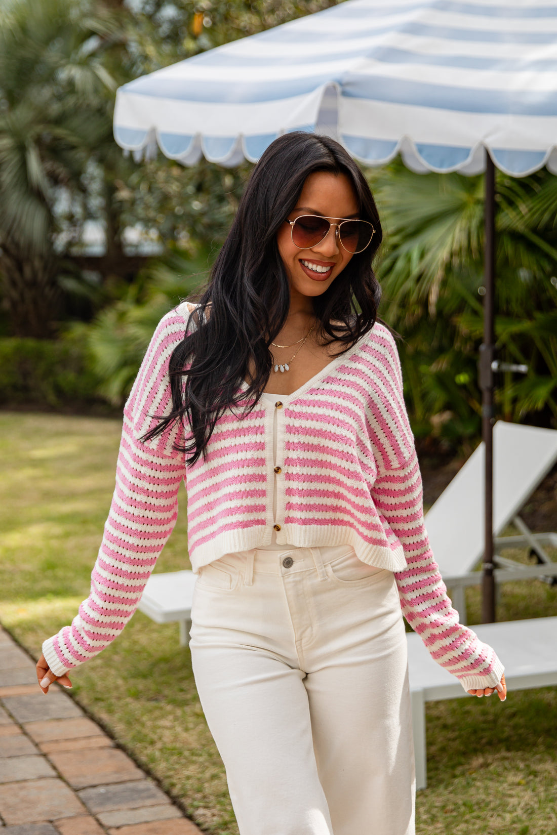 Woman wearing a pink and white striped sweater and sunglasses outdoors under a striped umbrella.