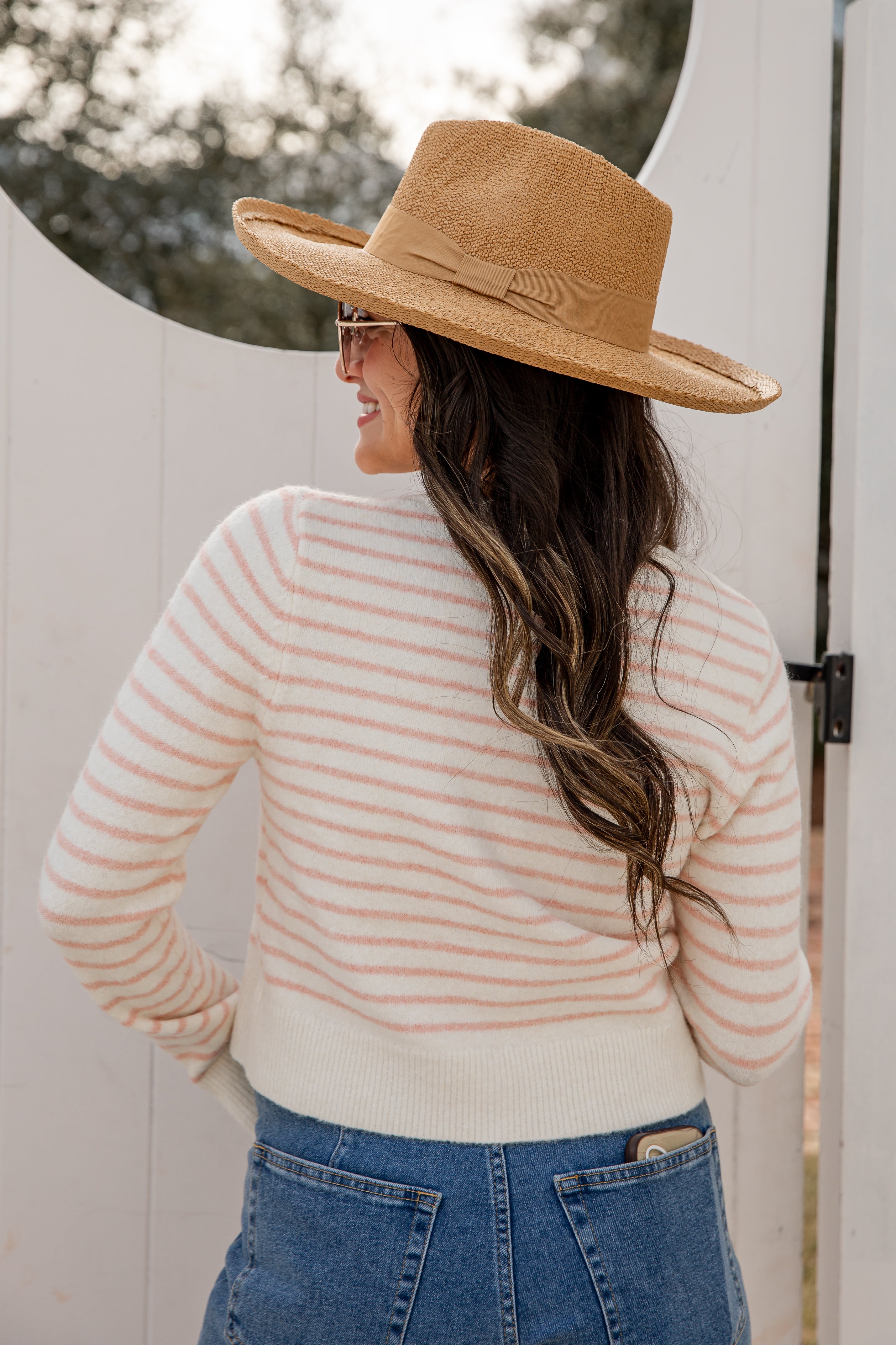 Woman wearing a beige and pink striped sweater, blue jeans, and a wide-brimmed brown hat.