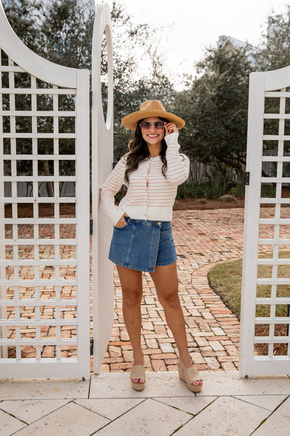 Woman standing behind a white lattice gate wearing a striped sweater, denim shorts, and a hat.